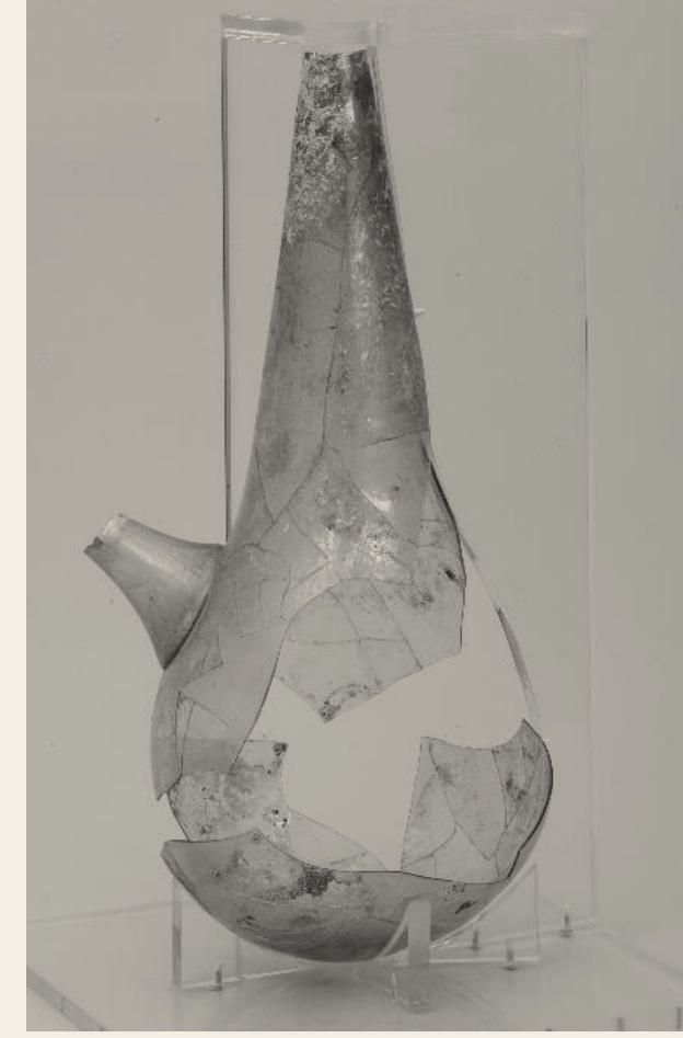 Photograph of a broken glass alembic with a side spout, mounted on a clear stand.