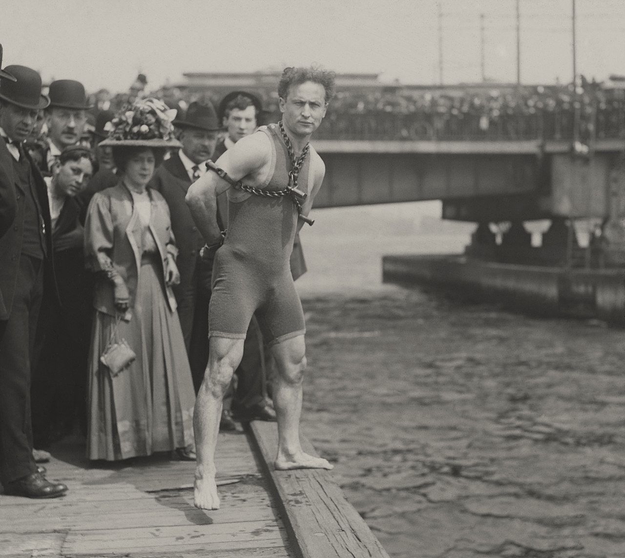 Harry Houdini in chains and a bathing suit on a pier beside onlookers.