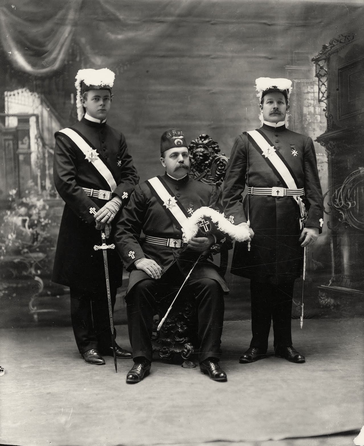 Studio photograph of three uniformed officers with plumed hats, sashes and swords, one seated.