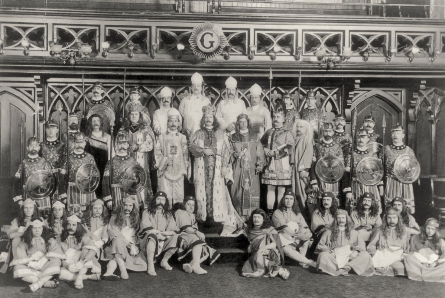 Group photograph of several rows of men in robes, crowns and costume before a carved screen.